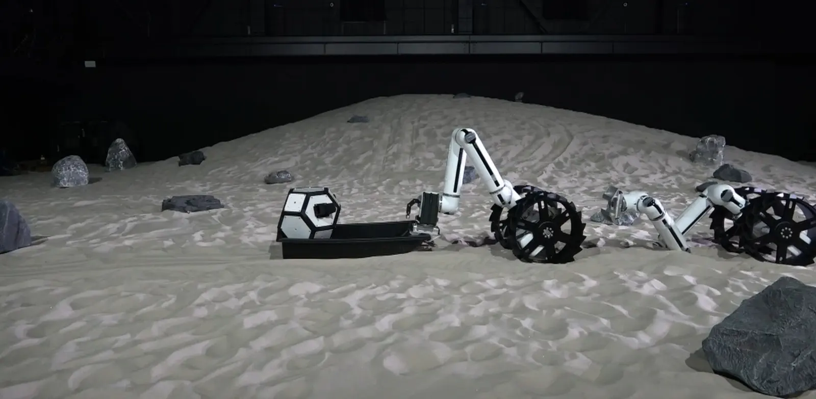 Two MoonBot configurations working as a team, a Minimal and a Dragon, clearing rocks to prepare the ground for infrastructure, JAXA Space Exploration Test Field.