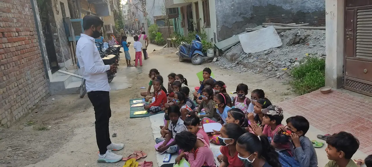 A volunteer teaching a class of children seated on mats in a narrow residential lane.